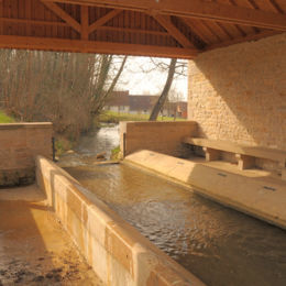 Ce lavoir couvert est aménagé à cheval sur un cours d'eau. Le bâtiment est composé de deux murs latéraux et d'une charpente en bois couverte en tuiles. Dans la partie protégée par le bâtiment, le fond du cours d'eau est dallé et il est bordé par une rangée de pierres inclinées sur chaque côté. Le cours d'eau est libre et aucun aménagement ne semble avoir été prévu pour régler la hauteur du bassin. La zone de lavage est surbaissée par rapport au niveau du cours d'eau pour permettre le lavage debout.
Chaque côté bénéficie d'un banc en pierre. Une dalle calcaire permet de traverser le cours d'eau pour accéder aux deux zones de lavage. Une rambarde métallique assure la sécurité du passage.
06/2017 F Leng phot. 06 caue39 - VINCELLES (39190)